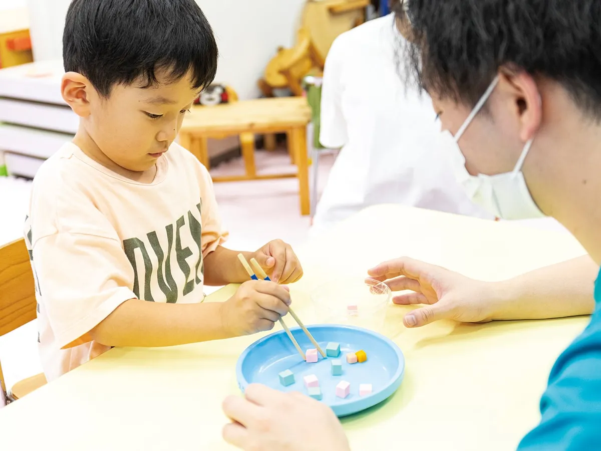 作業療法士に見守られ、箸で積み木をつまむ練習に取り組む子ども
